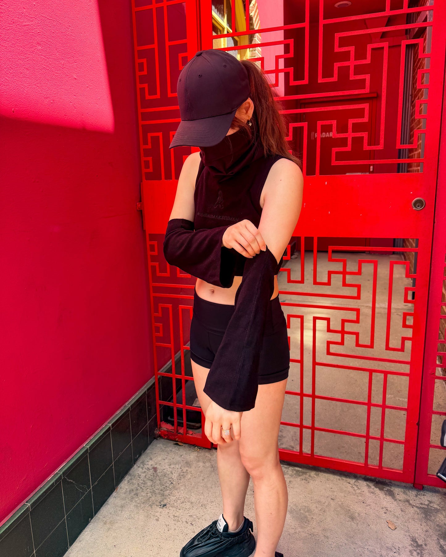 A person wearing a black sleeveless crop top, black shorts, a black baseball cap, and a black face mask stands in front of a bright red geometric metal gate and red wall.