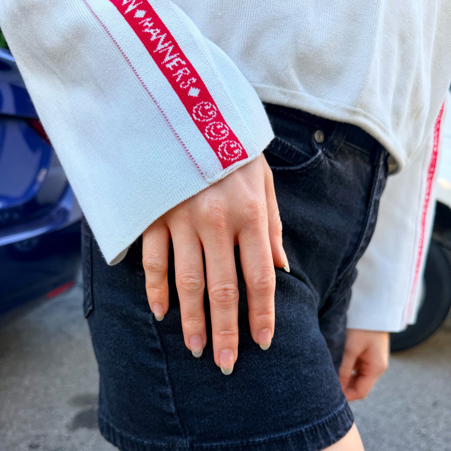 A close-up of a person wearing dark wash denim shorts and a white sweater with red stripes and smiley face details on the sleeves, standing outdoors on asphalt.
