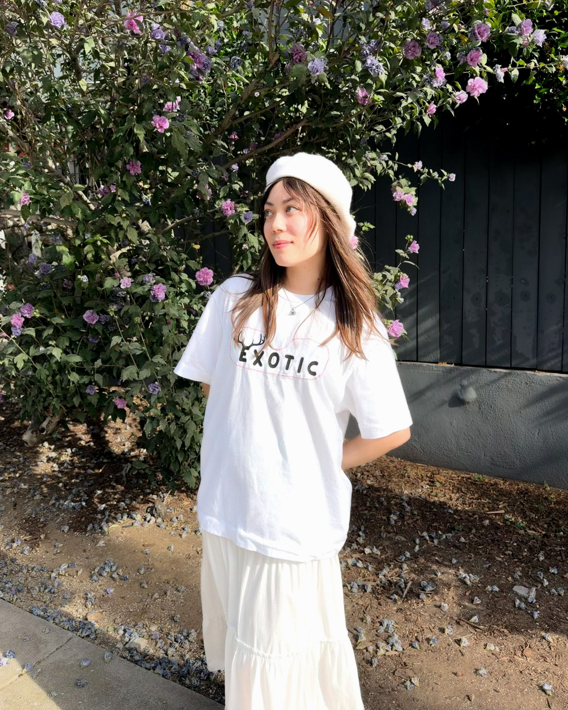 A woman wearing a white short-sleeve t-shirt with 'EXOTIC' printed in black across the chest, paired with a white long skirt and a white beret, standing outdoors in front of a flowering bush.