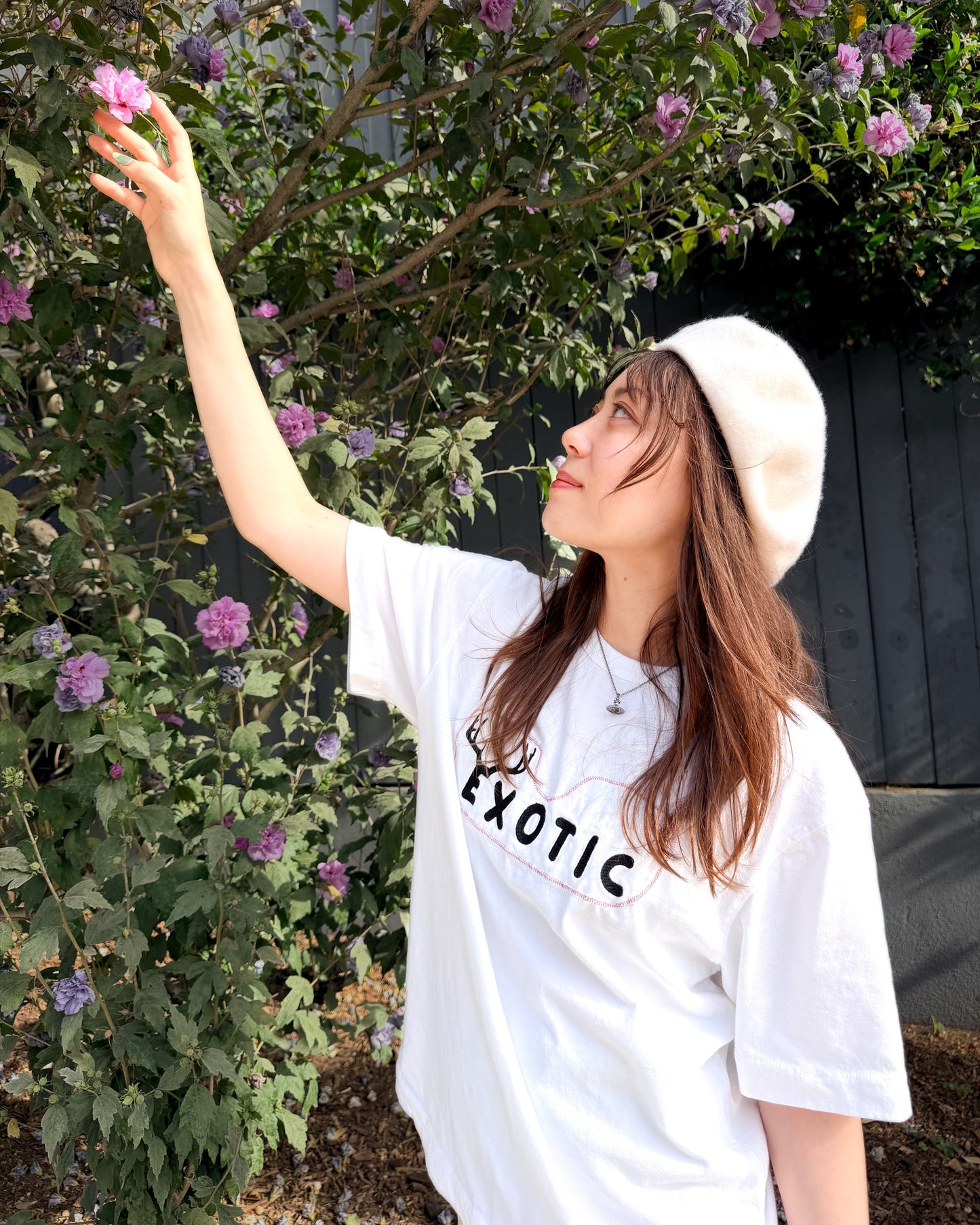 A woman wearing a white graphic t-shirt with 'EXOTIC' printed on the chest, a white beret, and a necklace, posing outdoors in front of a flowering bush with purple blossoms.