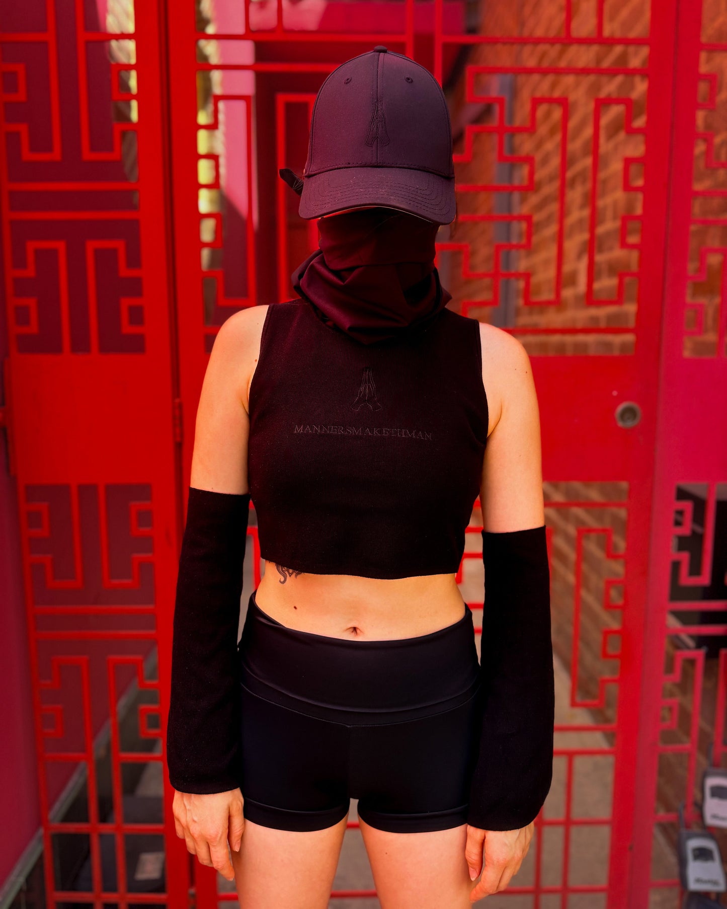 A person stands facing forward wearing a black sleeveless crop top, black arm warmers, black shorts, and a dark baseball cap in front of a bright red decorative screen.