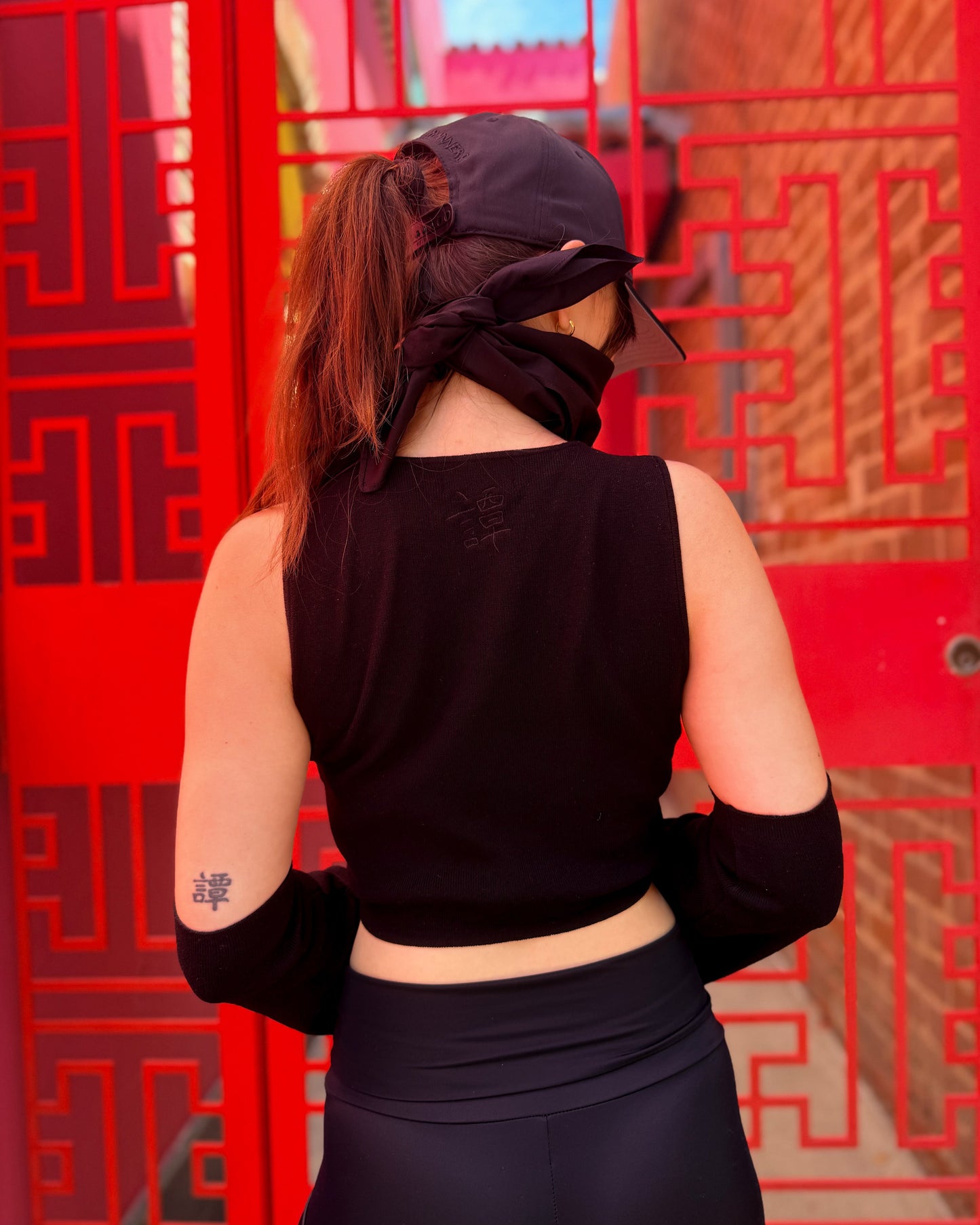A woman viewed from behind wearing a black sleeveless crop top, black leggings, a black cap, and a black face covering, standing in front of a bright red patterned door.