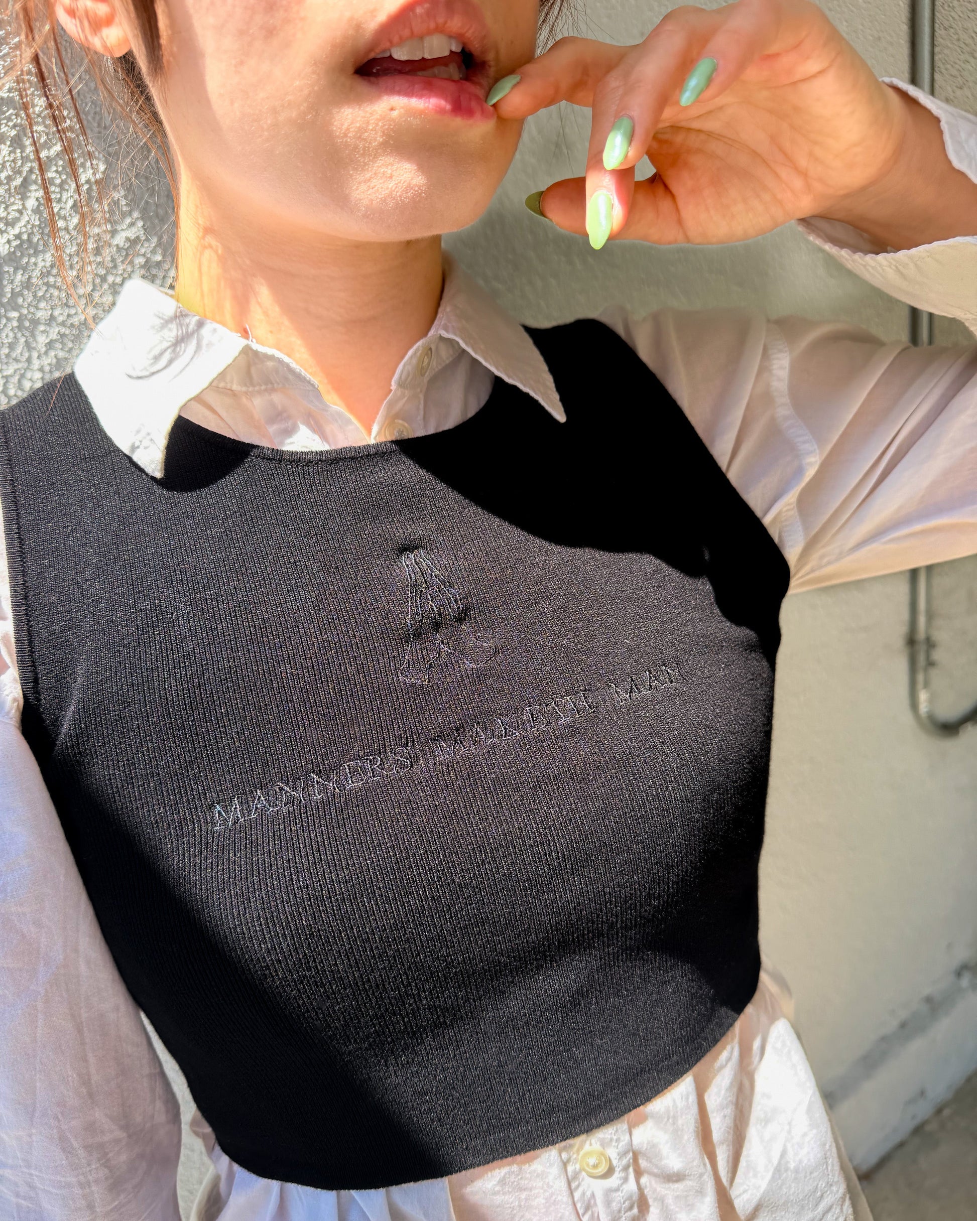 A close-up of a person wearing a black ribbed knit crop top with a white collared shirt underneath. The top features a tonal embroidered logo and text on the chest.