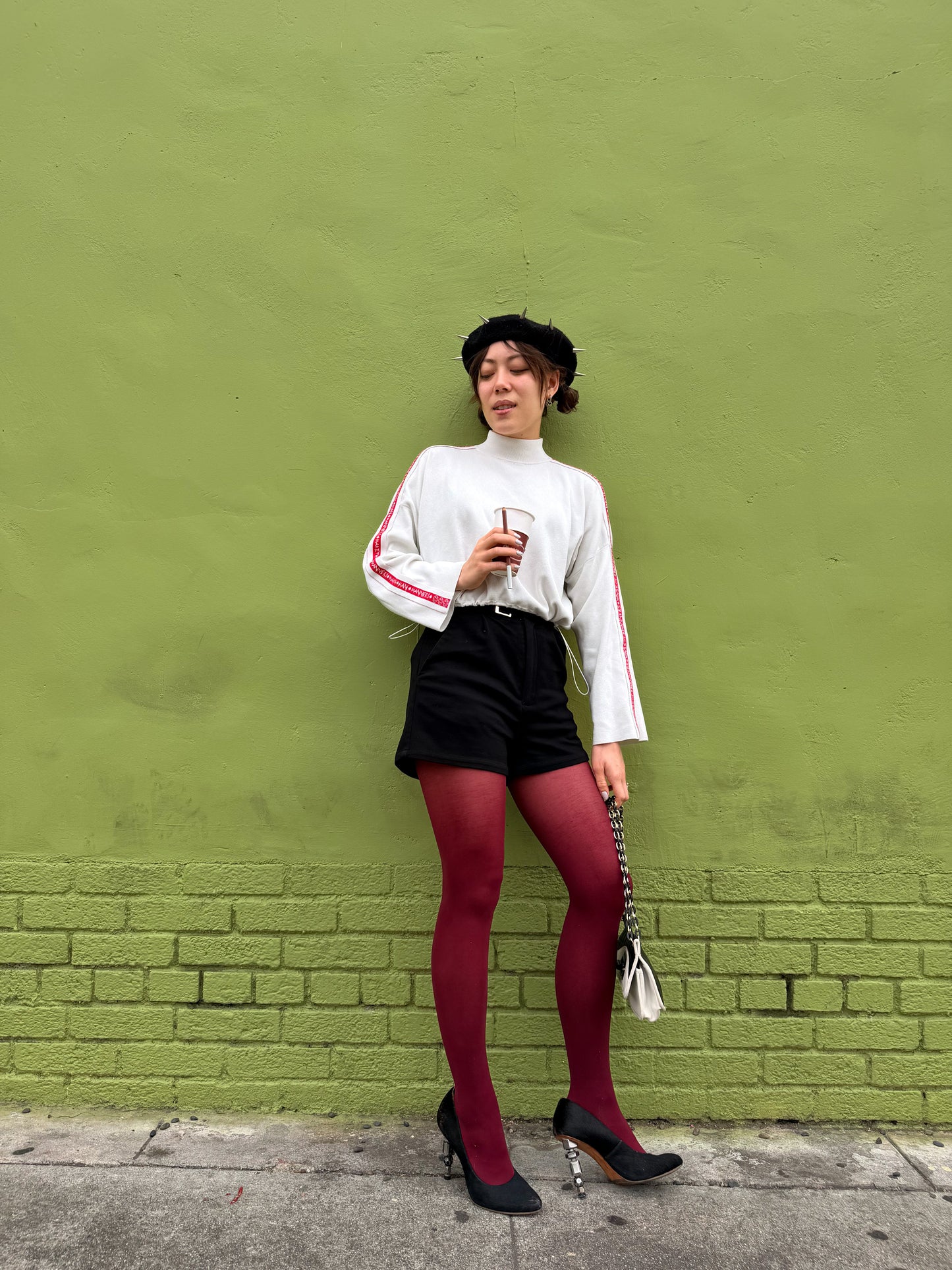 A woman stands against a textured green wall wearing a white mock-neck long-sleeve top with red stripes on the sleeves, black shorts, red tights, and black heels.