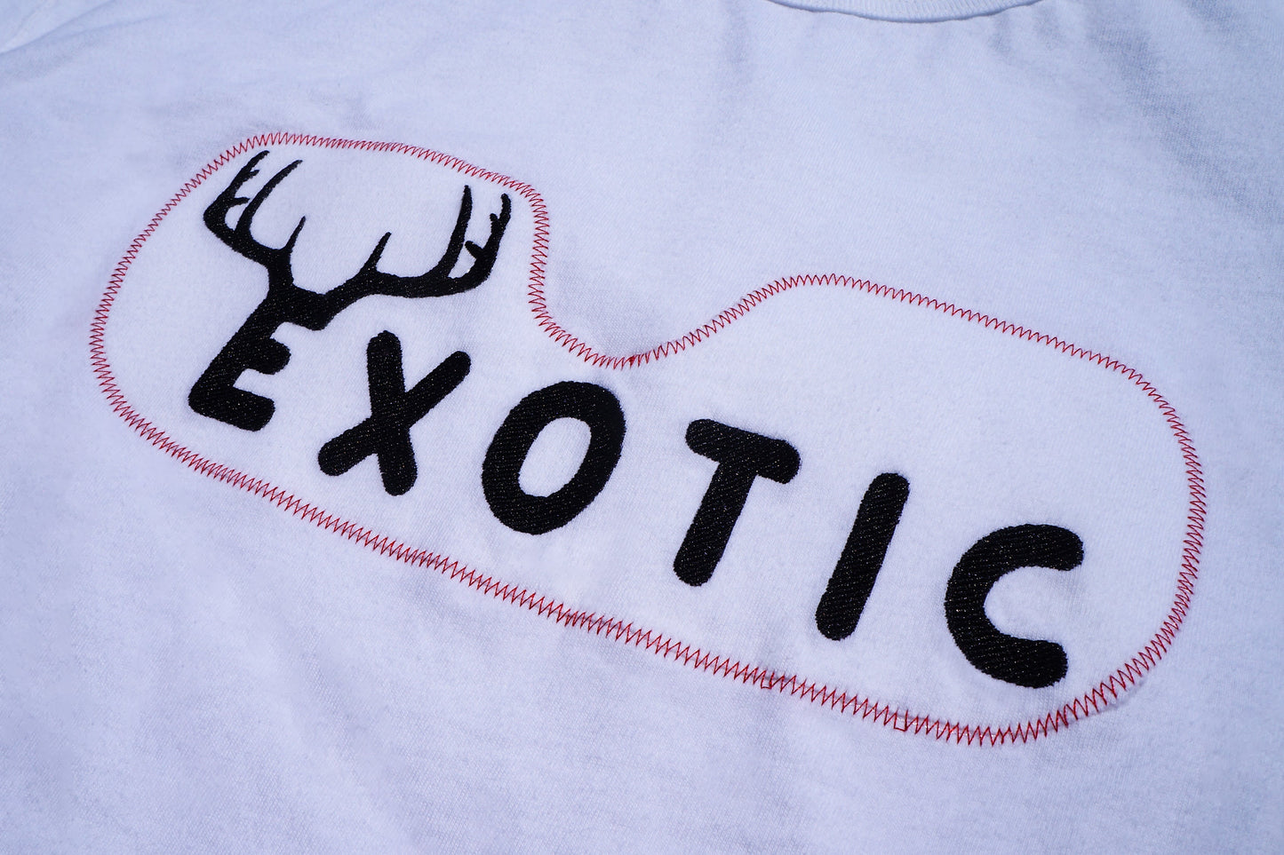 Close-up of a white fabric surface with a black embroidered wordmark reading "EXOTIC" inside a red stitched outline, plus a black antler graphic above the text.