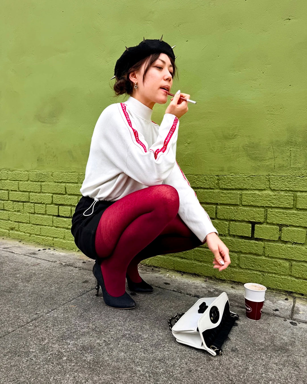 A woman crouches on a sidewalk against a green wall, wearing a white long-sleeve top with red trim, a black beret, black shorts, and dark red tights. A white handbag and a coffee cup sit on the ground beside her.