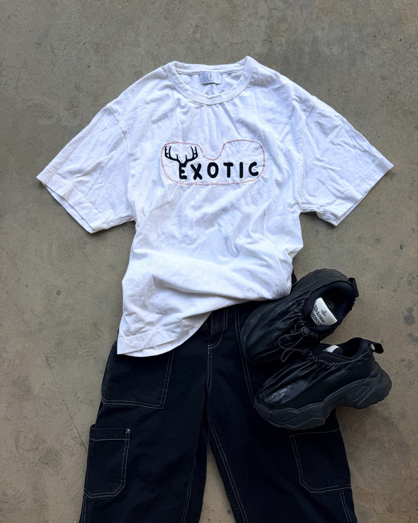 A white short-sleeve graphic T-shirt with a black deer antler and the word 'EXOTIC' printed on the chest, styled in a flat lay with black cargo pants and black sneakers on a concrete floor.