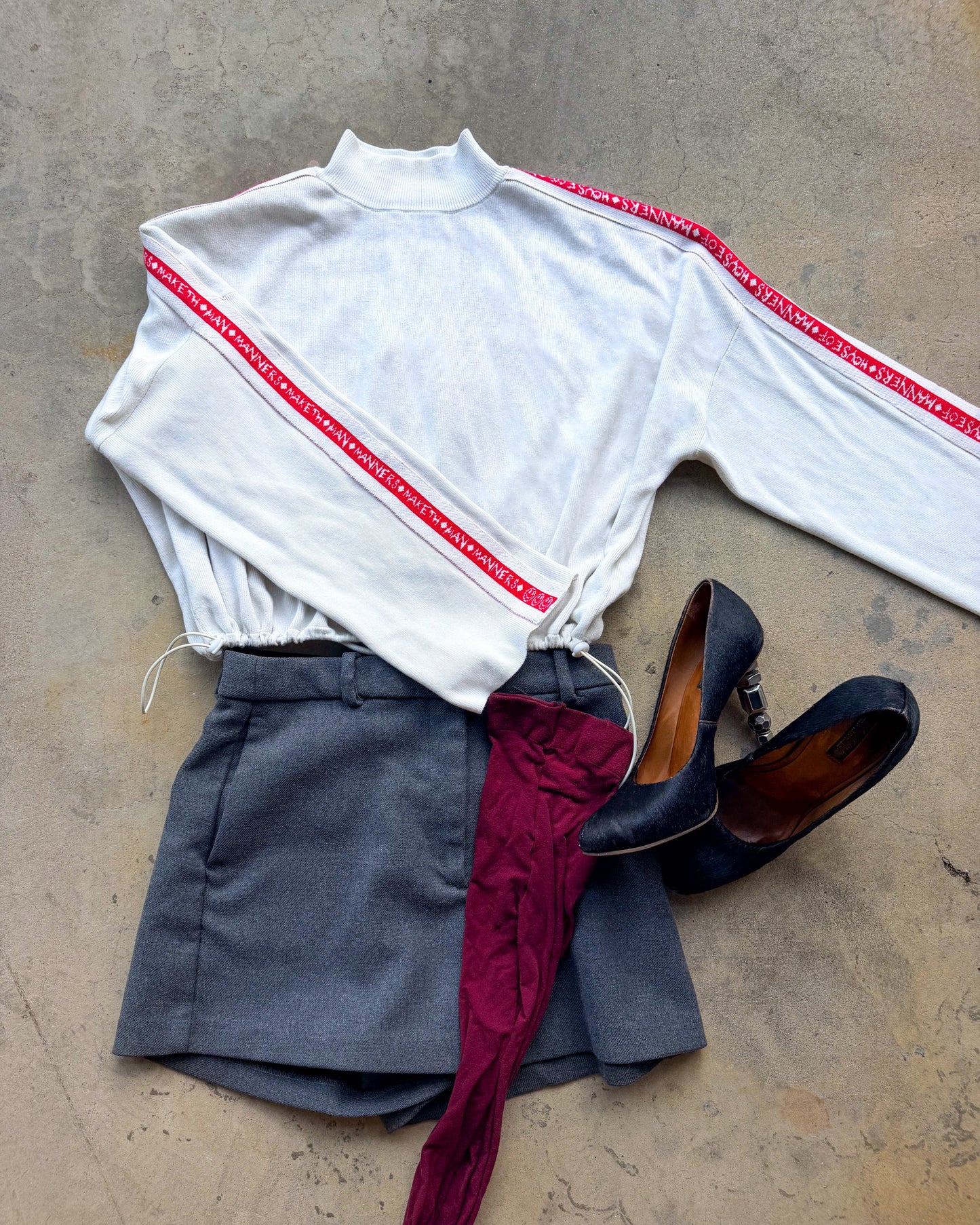A styled flat-lay outfit on a concrete floor featuring a white mock-neck sweatshirt with red stripes, gray shorts, burgundy tights draped over the shorts, and black pointed-toe heels.