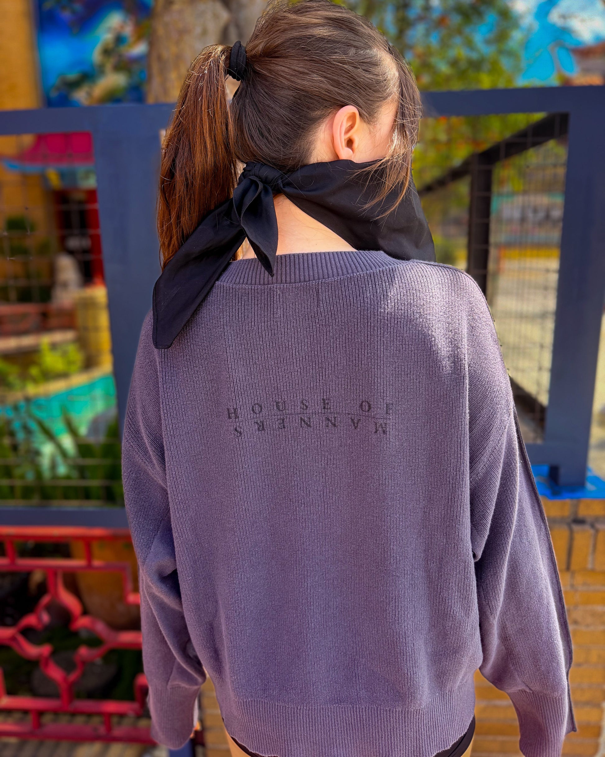 A woman viewed from behind wearing a blue-gray ribbed knit sweater with 'HOUSE OF MANNERS' printed in dark gray on the back, styled with a black neck scarf.