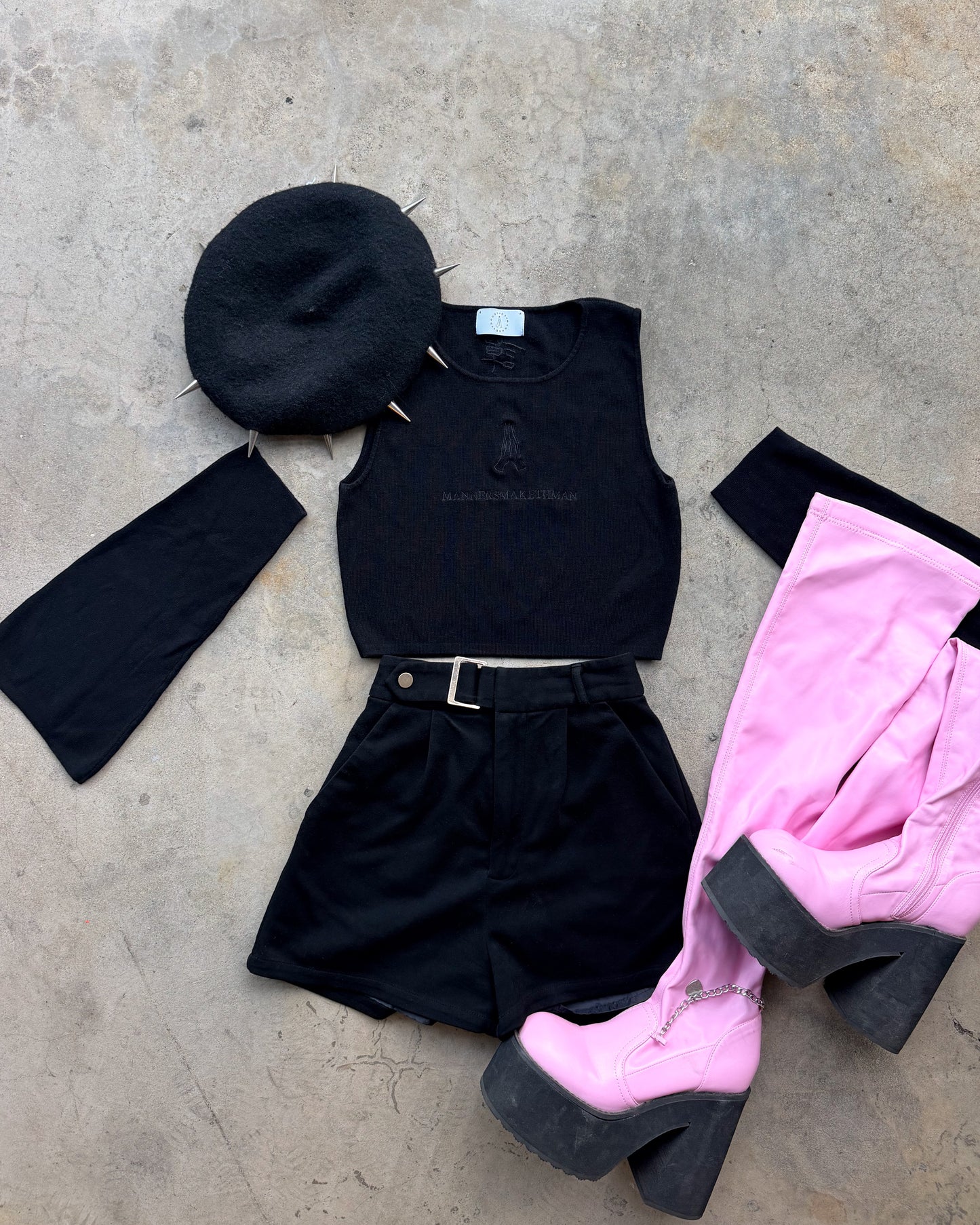 A styled flat-lay outfit on a concrete floor featuring a black studded beret, a black sleeveless top, black shorts with a belt, and a pair of bright pink platform high-heeled boots with black soles.
