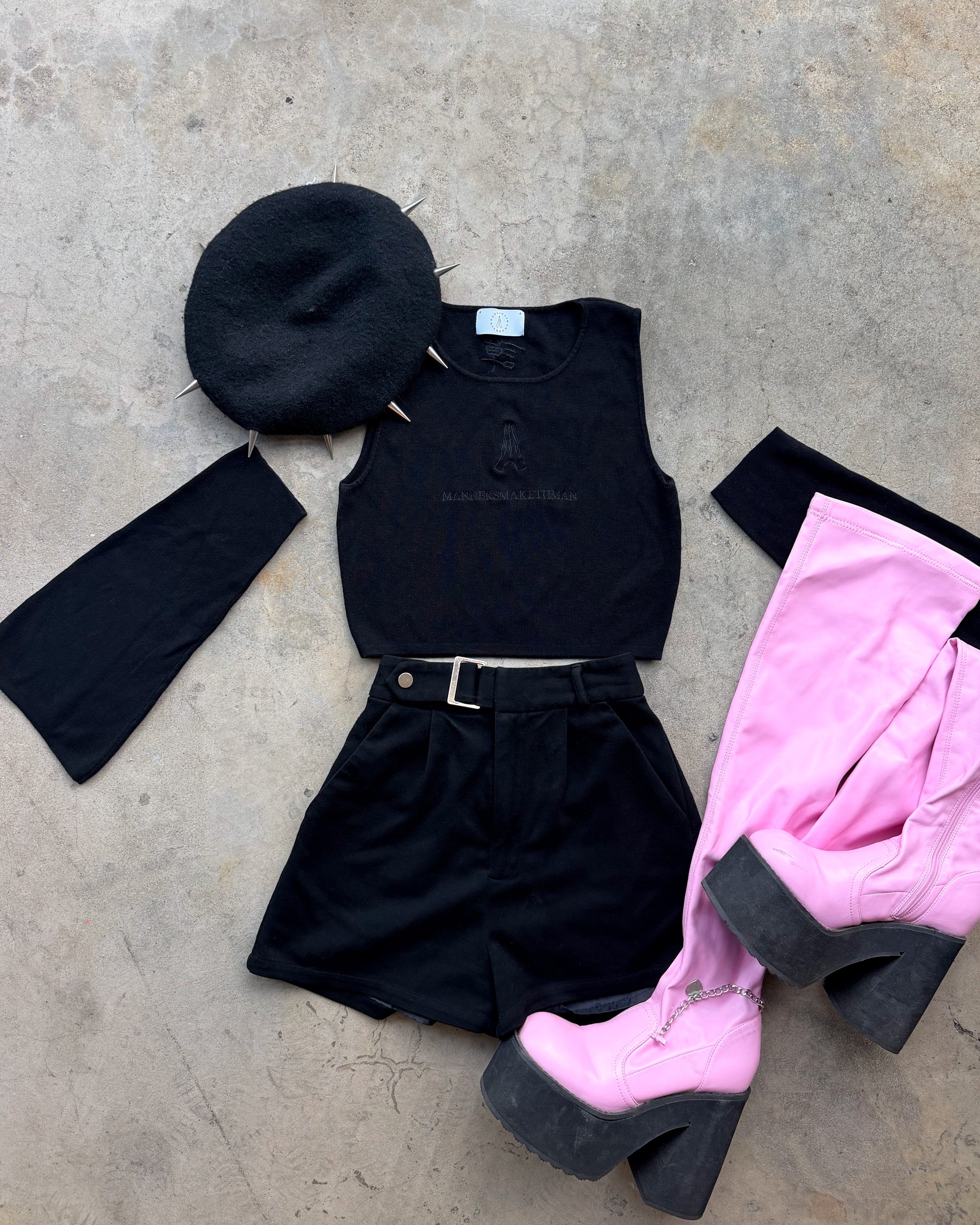 A styled flat-lay outfit on a concrete floor featuring a black studded beret, a black sleeveless top, black shorts with a belt, and a pair of bright pink platform high-heeled boots with black soles.